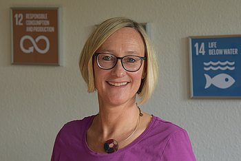 Eine Frau mit blonder Kurzhaarfrisur, die vor Schildern mit den Nachhaltigkeitszielen (SDGs) steht. Sie trägt eine violette Bluse und lächelt in die Kamera.
