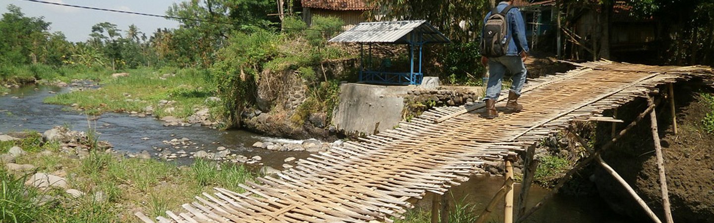 Eine einfache Holzbrücke über einen Fluss in einer ländlichen Landschaft in Indonesien.