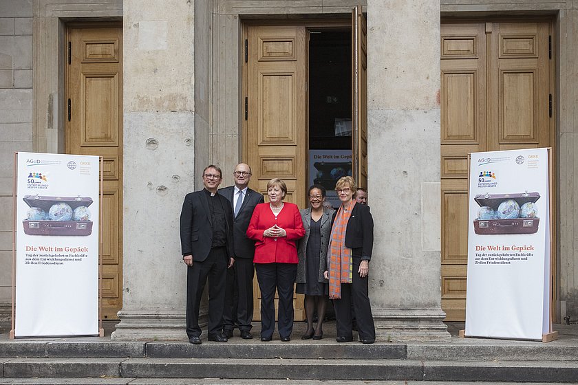 Eine Gruppe von fünf Personen steht auf den Stufen vor einem Gebäude mit großen Holztüren. Zwei weiße Roll-Up-Banner mit Bildern und Text flankieren den Eingang. Die Person in der Mitte trägt einen roten Blazer, der Hintergrund zeigt eine historische Architektur mit Säulen.