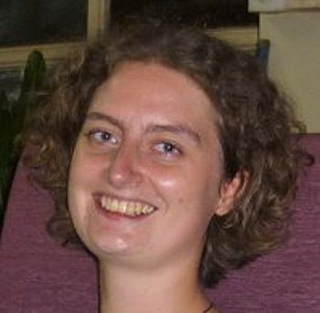 Porträt einer jungen Frau mit lockigem Haar und einem blauen Oberteil. Sie sitzt auf einem lila Stuhl und lächelt in die Kamera.