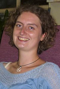 Porträt einer jungen Frau mit lockigem Haar und einem blauen Oberteil. Sie sitzt auf einem lila Stuhl und lächelt in die Kamera.