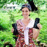Eine Frau steht im Freien und hält mehrere Pflanzen in schwarzen Behältern, lächelnd in die Kamera.