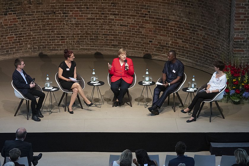 Fünf Personen sitzen auf einer Bühne in einem Podiumsgespräch, die Stühle sind in einem Halbkreis angeordnet. In der Mitte trägt eine Frau eine rote Jacke, umgeben von anderen Teilnehmenden in schwarzer und weißer Kleidung. Der Hintergrund zeigt eine Backsteinwand, während das Publikum im Vordergrund teilweise sichtbar ist.