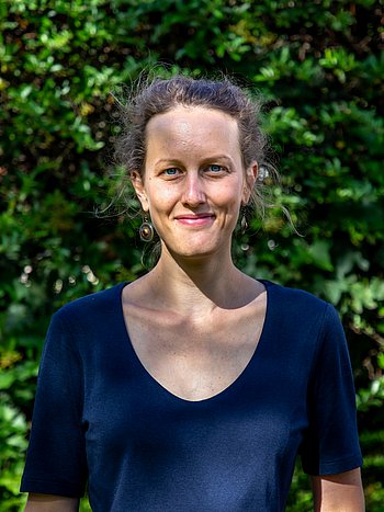 Eine Frau mit schulterlangem braunem Haar steht in einem Garten vor grünem Laub. Sie trägt ein dunkelblaues T-Shirt und lächelt leicht in die Kamera.