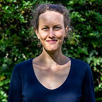 Eine Frau mit schulterlangem braunem Haar steht in einem Garten vor grünem Laub. Sie trägt ein dunkelblaues T-Shirt und lächelt leicht in die Kamera.
