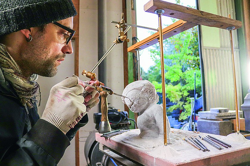 Ein Mann arbeitet konzentriert an einer kleinen Skulptur mit präzisem Werkzeug. Die Szene spielt in einem Atelier mit natürlichem Licht, und im Hintergrund ist ein Blick ins Grüne zu erkennen.