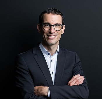 Ein mittelalter Mann mit schwarzen Haaren, Brille und schwarzem Anzug lächelt freundlich in die Kamera.