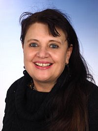 Porträt einer Frau mit dunklen, langen Haaren, die in die Kamera lächelt. Sie trägt einen schwarzen Schal und hat einen hellen Hintergrund.