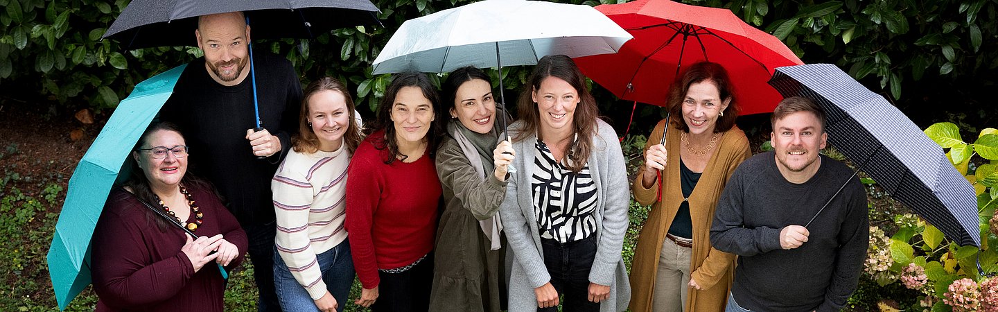 Gruppenfoto im Freien, bei dem mehrere Personen unter bunten Regenschirmen stehen. Im Hintergrund sind grüne Büsche und Blumen zu sehen. Die Gruppe lächelt in die Kamera, die Szene wirkt fröhlich und ungezwungen.