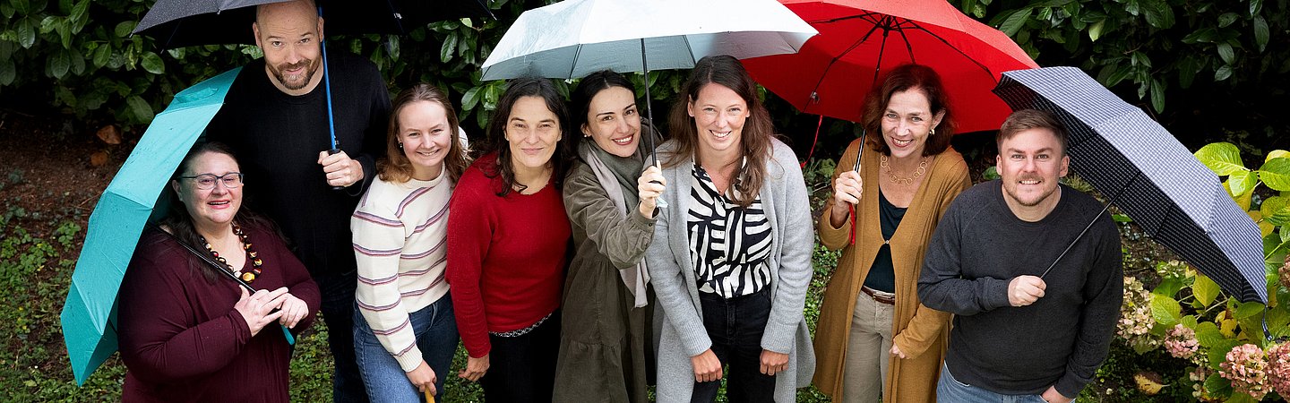 Gruppenfoto im Freien, bei dem mehrere Personen unter bunten Regenschirmen stehen. Im Hintergrund sind grüne Büsche und Blumen zu sehen. Die Gruppe lächelt in die Kamera, die Szene wirkt fröhlich und ungezwungen.