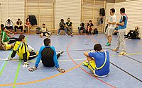 Kinder spielen in einer Turnhalle, während Erwachsene am Rand stehen und zuschauen.