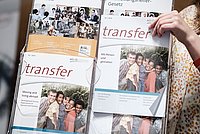 Eine Hand greift nach einer Ausgabe des Magazins 'Transfer', das in einem Ständer mit weiteren Exemplaren ausgestellt ist.