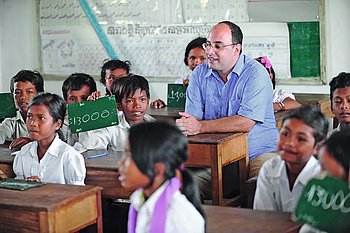 Ein Klassenzimmer mit Kindern, die an Holzbänken sitzen und kleine Tafeln halten. Eine Fachkraft  in einem blauen Hemd sitzt zwischen den Schülern und nimmt an der Unterrichtssituation teil. Der Raum ist einfach eingerichtet und hell.