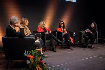 Ein Podiumsgespräch mit fünf Personen, die auf einer Bühne sitzen und miteinander diskutieren. Blumenarrangements schmücken die Bühne.