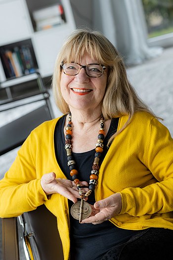 Eine Frau sitzt an einem Tisch, umgeben von Büchern und Schreibmaterialien, und lächelt in die Kamera.