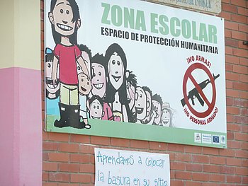 Ein Schild an einer Schulmauer mit der Aufschrift „Zona Escolar – Espacio de Protección Neutral“ und einer Illustration von Kindern, das ein Schutzgebiet für Kinder markiert.
