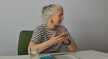 Zwei Frauen sitzen sich gegenüber. Hinter ihnen eine Wand mit der Aufschrift "UN VOLUNTEERS: We are inspiration in action".