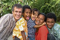Lachende Gruppe von Kindern und einem Erwachsenen vor grüner Pflanzenkulisse.