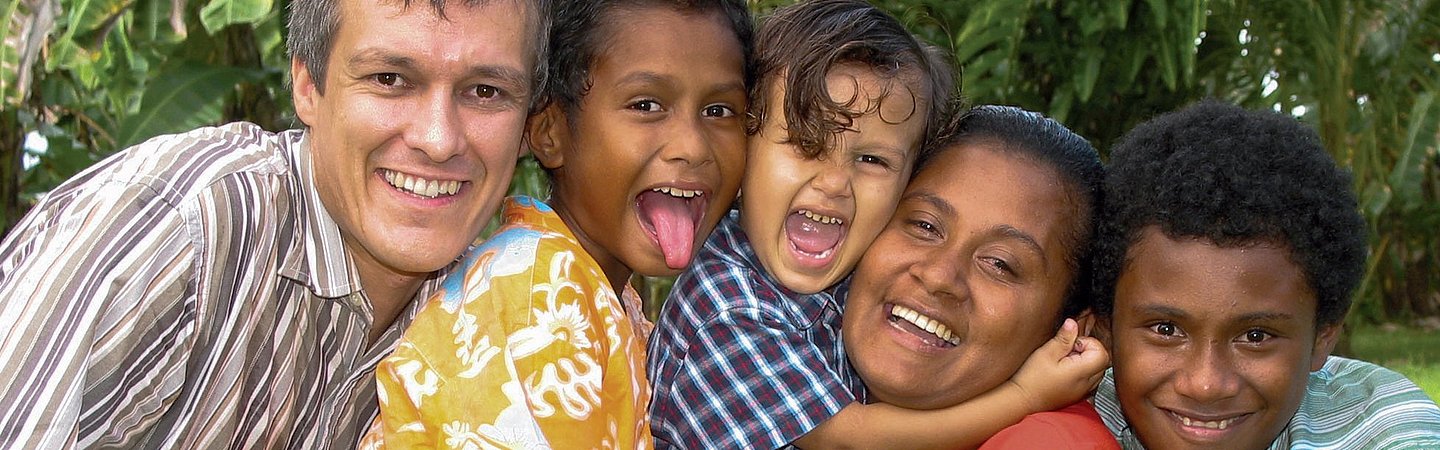 Lachende Gruppe von Kindern und einem Erwachsenen vor grüner Pflanzenkulisse.