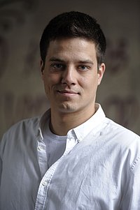 Porträt eines Mannes mit kurzem dunklem Haar, der ein weißes Hemd trägt. Der Hintergrund ist dezent und dunkel gehalten.