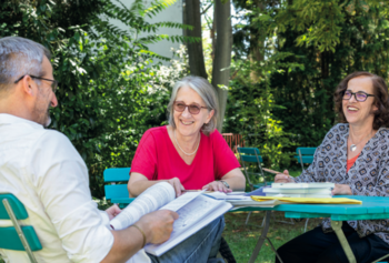 Drei Personen sitzen an einem Tisch im Freien, umgeben von grüner Vegetation. Eine Frau in einem roten Oberteil lächelt, während sie mit den anderen diskutiert. Auf dem Tisch liegen Notizbücher und Unterlagen, die eine entspannte Arbeitsatmosphäre im Garten andeuten.