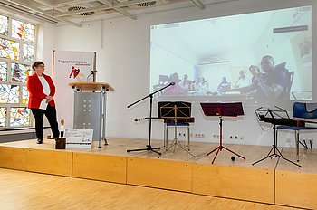 Eine Frau in einem roten Blazer steht auf einer Bühne neben einem Rednerpult und hält eine Präsentation. Im Hintergrund ist eine große Leinwand mit projizierten Bildern zu sehen. Der Raum ist hell und modern gestaltet, mit großen Fenstern, die Tageslicht hereinlassen.