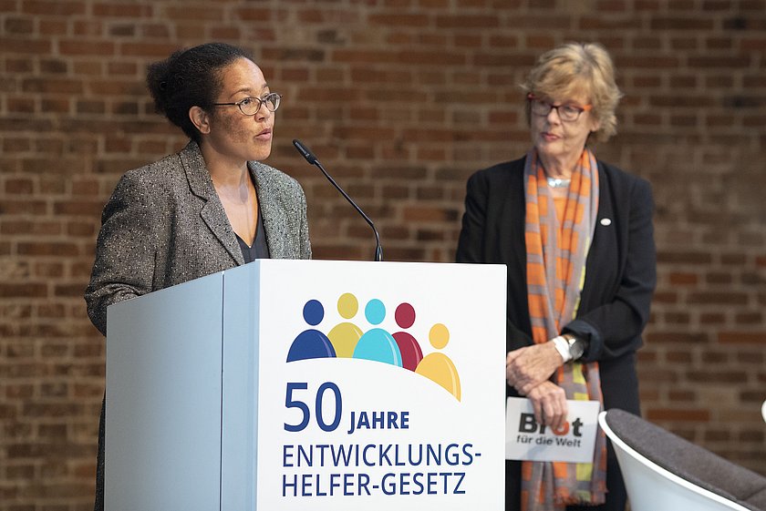 Zwei Frauen stehen an einem Rednerpult mit der Aufschrift '50 Jahre Entwicklungshelfer-Gesetz'. Die linke Frau spricht ins Mikrofon und trägt eine gemusterte Jacke, während die rechte Frau mit einer Brille und einem orangefarbenen Halstuch aufmerksam zuhört. Im Hintergrund ist eine Backsteinwand zu sehen.