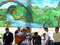 Eine Gruppe von Menschen spielt Musik im Freien vor einem großen, farbenfrohen Wandgemälde mit einer Landschaft, einem Regenbogen und einer Naturkulisse.