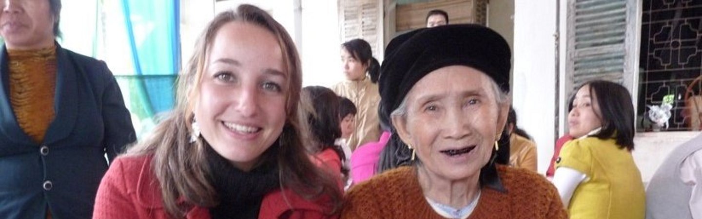 Zwei Frauen sitzen nebeneinander auf blauen Stühlen, eine von ihnen trägt eine rote Jacke, die andere ein orangefarbenes Outfit. Beide lächeln freundlich in die Kamera.